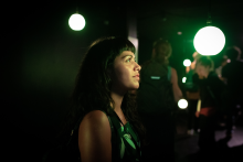 An image of a person with long black hair and a short fringe looking into the distance in a dark room, they are lit up in green neon light, and there are bright light bulbs scattered around in the background 